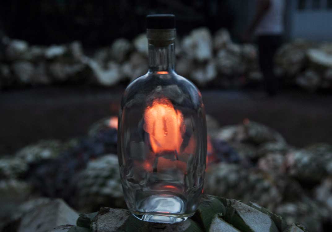 Botella de TLEM sin etiquetas y en el fondo el horno con las piñas y el fuego vivo